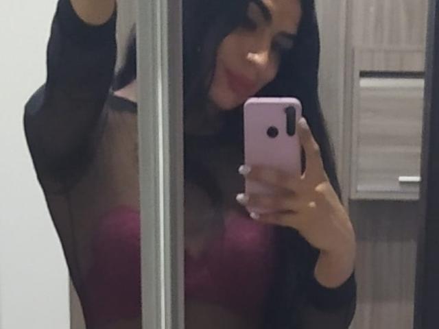 MikeyllaFoxT, con lunghi capelli neri, indossa una maglia nera trasparente e un reggiseno rosa, e sta scattando un selfie allo specchio con un telefono rosa, mostrando il seno..
