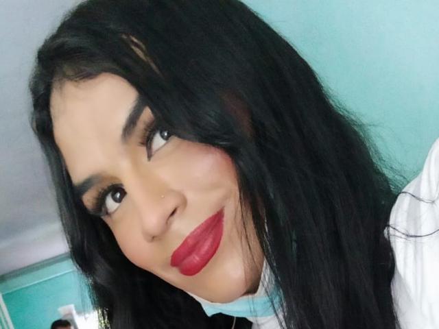 MikeyllaFoxT ha lunghi capelli neri, sopracciglia scure, rossetto rosso e un piccolo piercing al naso. Sta sorridendo con le labbra rosse e guarda leggermente di lato..