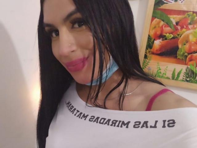 MikeyllaFoxT has long black hair, pink lipstick, and a blue mask, wearing a white shirt with a pink strap, smiling at the camera. Her tits are small.