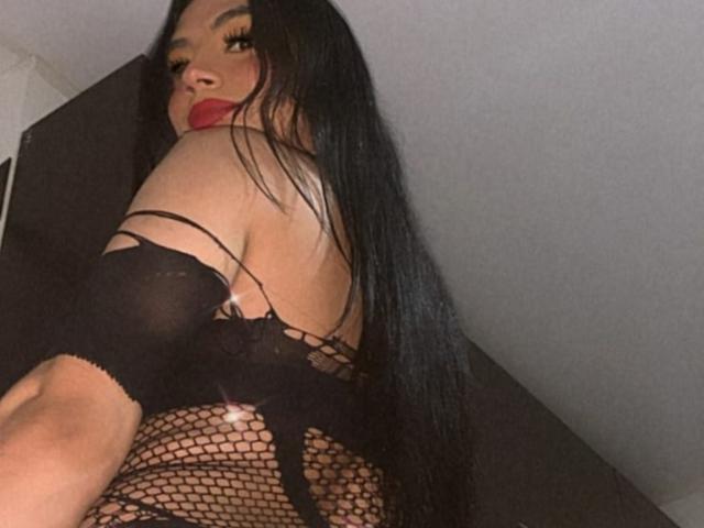 MikeyllaFoxT con lunghi capelli neri, rossetto rosso, lingerie in rete nera e schiena scoperta, che mostra glutei e schiena, mentre si guarda sopra la spalla..