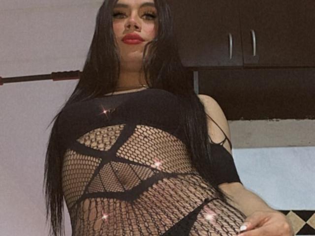 MikeyllaFoxT, con lunghi capelli neri, indossa un abito nero in rete a maglie con motivi geometrici, che mette in mostra il seno, ed è in piedi in una cucina, guardando verso l'alto con il rossetto rosso..