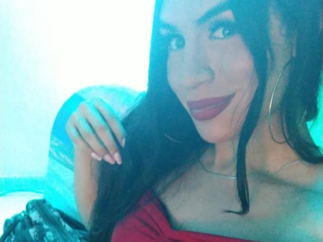MikeyllaFoxT with long dark hair, red lipstick, and hoop earrings, smiling at the camera, wearing a red top, showing her cleavage, and holding her hair.