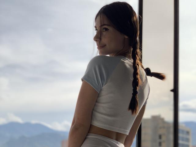 NikoleFoster with braided brown hair, white crop top, and matching white bottoms, looking over shoulder, smiling, standing by window, sunlight highlighting her ass, mountains in background.