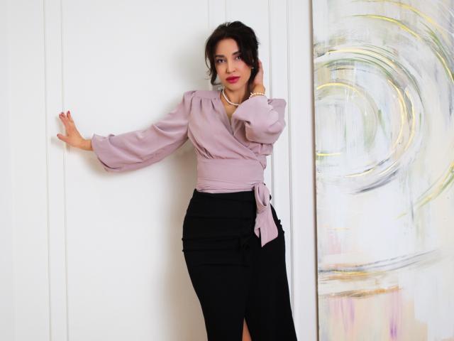 SimoniGrace, com uma blusa roxa volumosa, saia preta e colar, apoiada em uma parede branca, mão direita levantada, mão esquerda para baixo, sorriso discreto, batom vermelho, cabelo cacheado escuro, elegante..