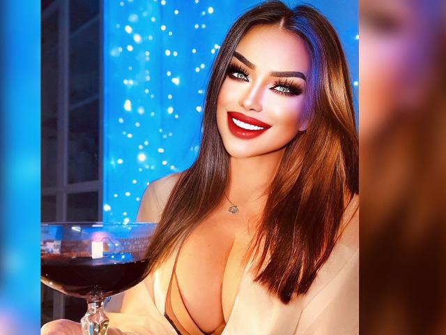 OhYeahh, com cabelos castanhos longos, seios grandes e batom vermelho, sorrindo para a câmera, segurando um copo de vinho tinto, vestindo uma blusa bege, em um ambiente com iluminação azul..
