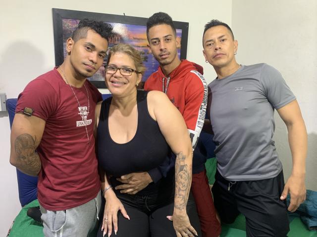 Four HottestFoursome standing in a room. HottestFoursome in front has big tits, black tank top, and glasses. HottestFoursome in red tracksuit, HottestFoursome in gray shirt, HottestFoursome with tattoos.