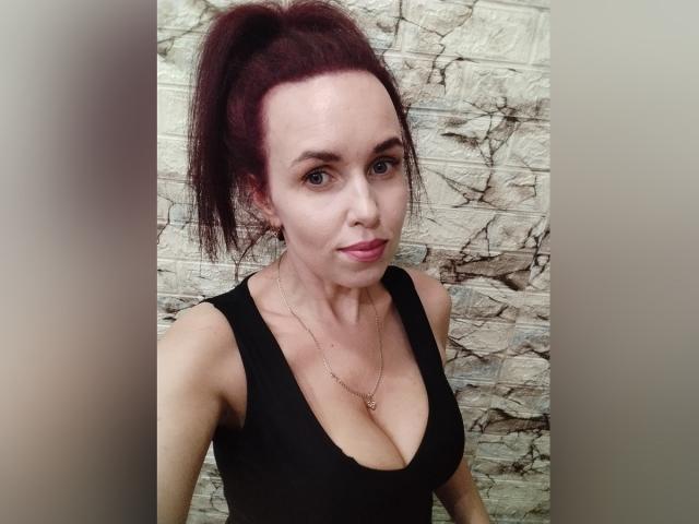 Allure, con una coleta roja, un top negro que deja ver sus grandes senos, piel clara, una nariz pequeña, labios rosados, y llevando un collar, frente a una pared de piedra con textura..