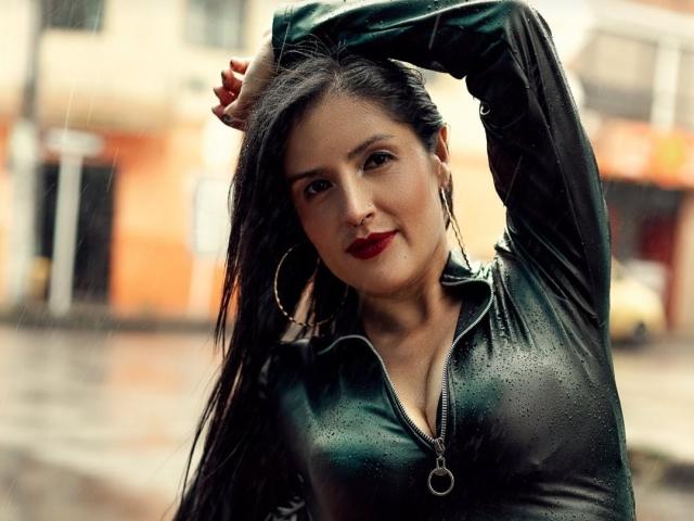 Adafieldd ha i capelli scuri, il rossetto rosso e indossa una giacca di pelle nera con cerniera e decorazioni a goccia. Ha un braccio alzato, indossa grandi orecchini a cerchio ed è leggermente sorridente..