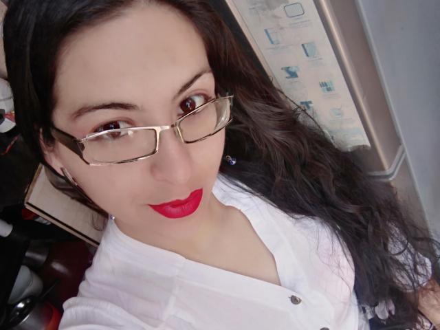 SashaGreish, con lunghi capelli neri, che indossa occhiali, rossetto rosso e una camicia bianca, guarda verso l'alto con un leggero sorriso, mostrando il naso e la parte superiore del petto. Il seno non è visibile..