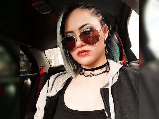 CassieJones with pale skin, black and green dyed hair, wearing oversized sunglasses, black choker, gray and black hoodie, and black tank top, sitting in a car.