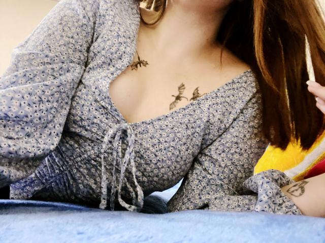 Rubbi with brown hair, wearing a blue floral dress, showing small tits with black leaf tattoos above, lying on a blue surface, holding a cigarette.