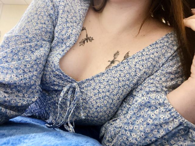 Rubbi has small tits with two butterfly tattoos above her cleavage, wearing a blue floral dress with a low neckline, her brown hair partially visible.