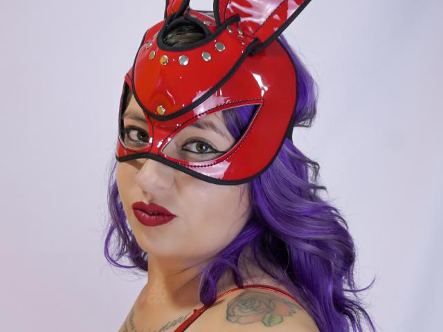 Nicoleks with purple hair, wearing a shiny red latex mask with black and gold studs, red lipstick, tattoos on her shoulder, and a red top, looking at the camera.