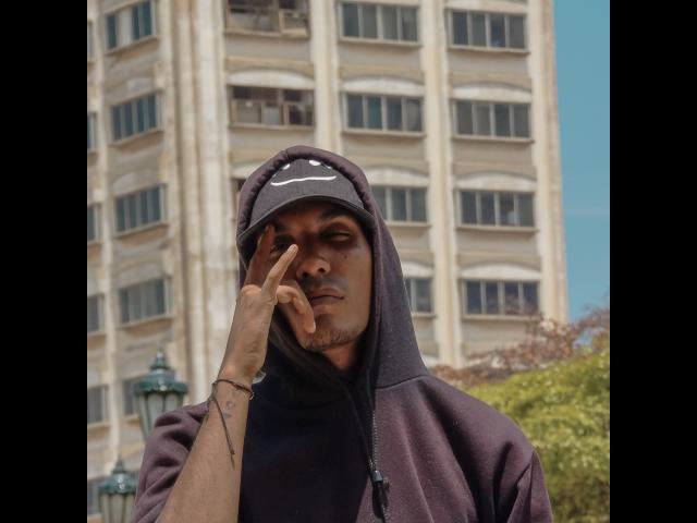 Shilver, un homme à la peau foncée avec une barbe de style bouc, portant un sweat à capuche et une casquette noirs Nike, touchant son visage avec un doigt, se tenant devant un grand bâtiment beige..