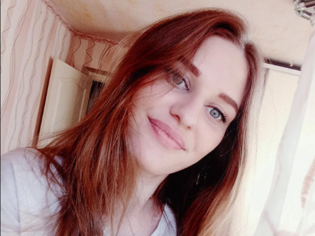 SanataDemori, con lunghi capelli rossi, occhi azzurri e pelle chiara, sorride leggermente, indossando una maglietta bianca, in una stanza luminosa. Ha il seno piccolo ed è molto carina..