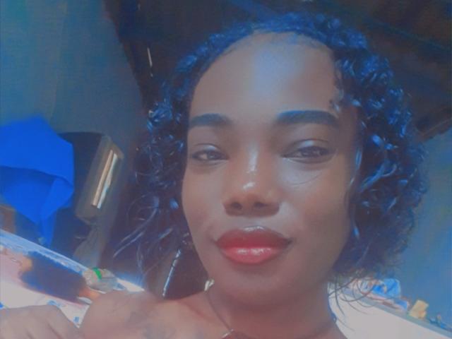 Shere has dark curly blue hair, shiny red lips, and a smooth black face, smiling at the camera with a small tattoo under her right eyebrow.