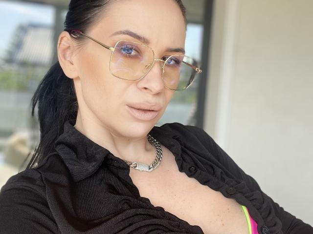 SabrinaMore with blue eyes, black shirt, gold glasses, and chain necklace, showing her tits in a pink bra, has dark hair in a ponytail, and a subtle smile.