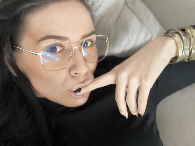 SabrinaMore with black turtleneck, gold glasses, finger in mouth, blue reflection on glasses, gold bracelets, black hair, light skin, and open mouth.