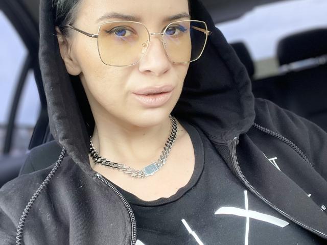 SabrinaMore, mit heller Haut, trägt eine schwarze Kapuzenjacke, eine Brille und eine silberne Kette. Sie blickt neutral in die Kamera. Die Brust ist nicht sichtbar..