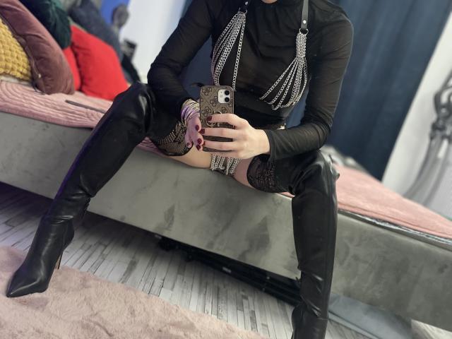 SabrinaMore está sentada em uma cama, vestindo uma blusa preta transparente, correntes, botas pretas que vão até a coxa e meias de renda, tirando uma selfie no espelho com o celular, mostrando seus seios..
