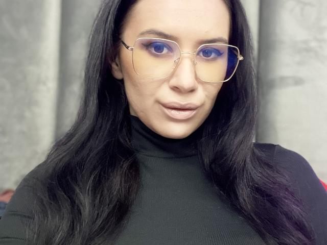 SabrinaMore, mit langem, schwarzem Haar, trägt eine goldfarbene Brille und einen schwarzen Rollkragenpullover. Sie hat blauen Lidschatten und leicht geschminkte, rosa Lippen und blickt direkt in die Kamera..