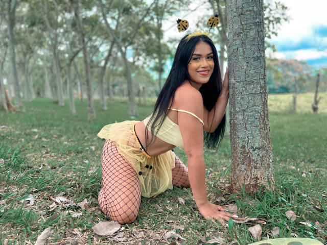 AlexaTorieli, agenouillée sur l'herbe, portant de la lingerie jaune, des bas résille et des oreilles en forme d'abeille, souriante, les mains sur un arbre, cheveux noirs longs, silhouette pulpeuse, poitrine moyenne, peau bronzée, fond de forêt..