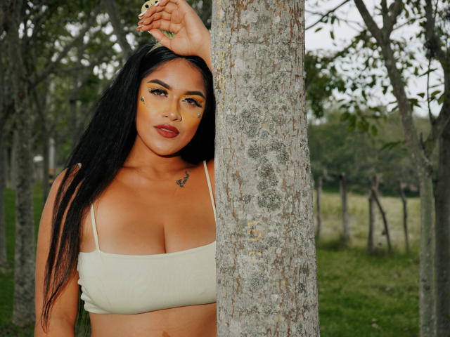 Alexa Torieli, con lunghi capelli neri, pelle abbronzata e trucco giallo sul viso, appoggiata a un albero, indossa un reggiseno verde, mostrando il suo seno prosperoso e un piccolo tatuaggio sul petto..