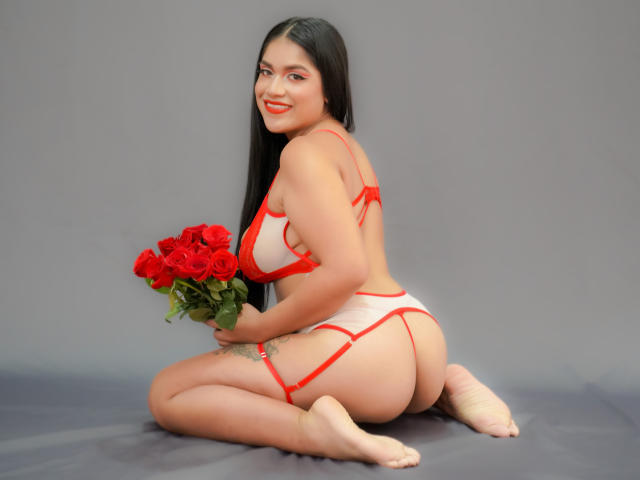 AlexaTorieli, met lang zwart haar, draagt rode lingerie, knielt op de grijze vloer, houdt rode rozen vast, glimlacht, toont haar billen en borsten, en kijkt over haar schouder..