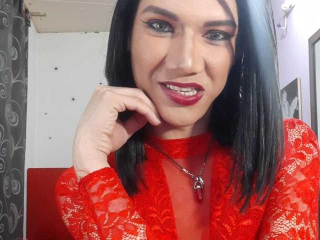 LunaGransTs met een rode kanten top, zwart haar en rode lipstick, lachend naar de camera, hand op de wang, ketting met een rode steen. Borsten zichtbaar door het kant..
