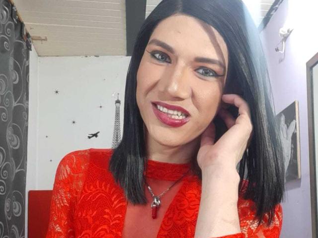 LunaGransTs con un corte de pelo bob negro, una blusa roja de encaje, lápiz labial rojo, sonriendo, con la mano en el pelo, ojos azules, un collar rojo, piel pálida, mejillas ligeramente sonrojadas, en una habitación bien iluminada..