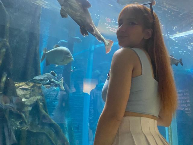 Melissa Skelton, avec de longs cheveux roux, un débardeur blanc et une jupe blanche, se tient dans un aquarium, tournée vers l'arrière, entourée de poissons, avec un dauphin au-dessus d'elle..