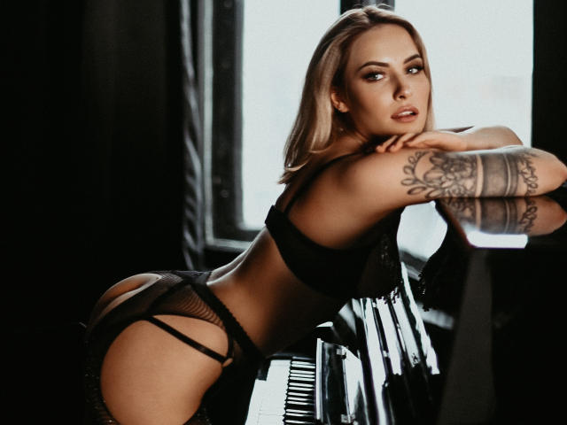 SuaSweetBlondie em lingerie preta, apoiada em um piano, mostrando o bumbum, o braço tatuado e olhando sedutivamente para a câmera, com cabelo loiro e uma expressão sexy e confiante..