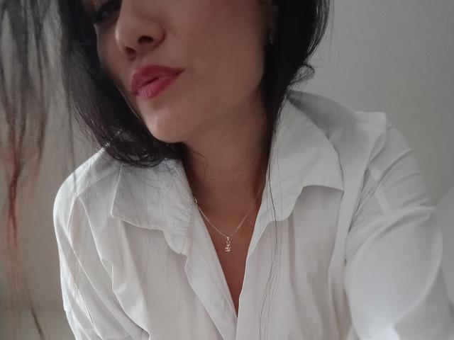 XBellaEvaX has dark hair, pink lipstick, and a white button-up shirt, with a small necklace, showing a slight smile, and her tits are partially visible.