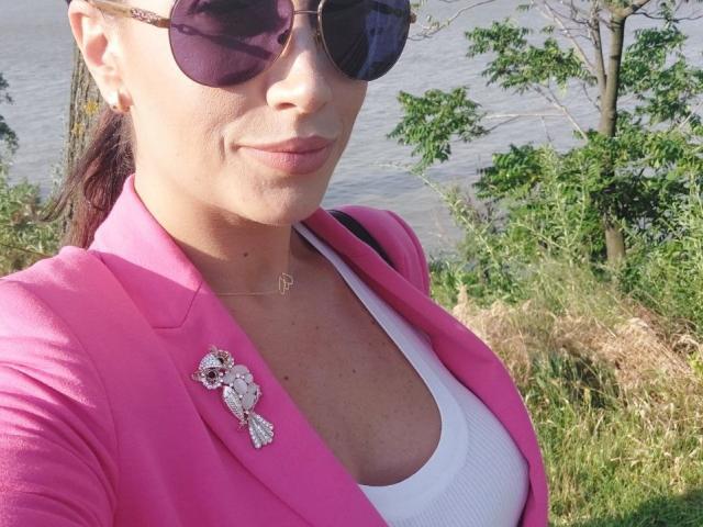 StassiLyanne portant un blazer rose, un haut blanc et une broche en forme de hibou rose, avec des lunettes de soleil foncées, montrant sa poitrine, un arrière-plan de feuilles vertes, de l'eau visible, des lèvres roses, une boucle d'oreille, un collier et un léger sourire..