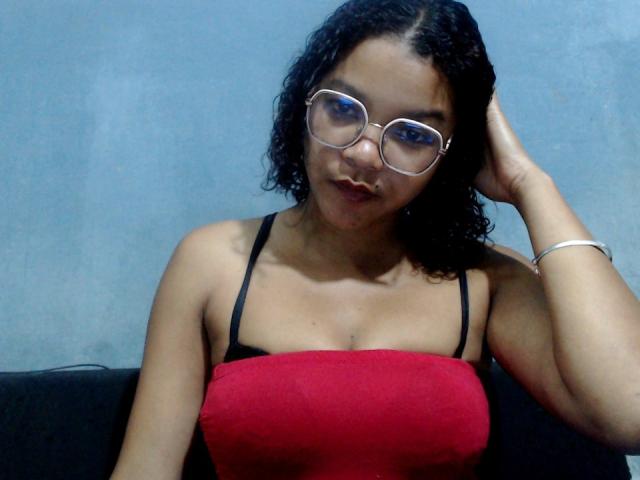 IreneSexy69 with curly black hair, glasses, and red top, showing her tits, black bra straps, and hand in her hair, looking at the camera with a slight smile.