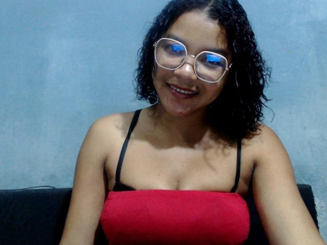 IreneSexy69, con cabello negro rizado, usando gafas y un top rojo sin tirantes, mostrando sus pequeños senos y sonriendo a la cámara. Su piel morena es visible..