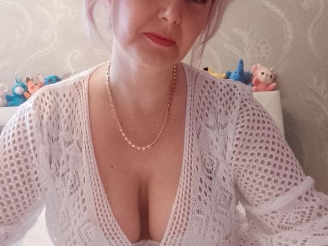 VAlica with pale skin, pink hair, and red lipstick, wearing a white, deep V, knit top, showing her boobs, and a pink beaded necklace, sitting in a bedroom.