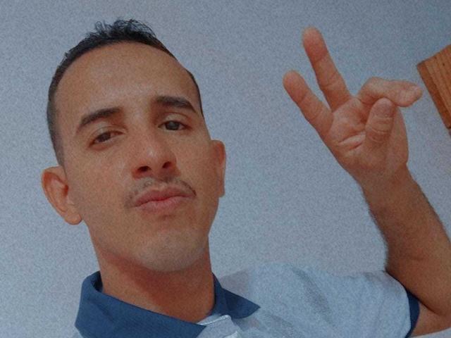 A photograph of Avel with short dark hair, a small mustache, and a light skin tone. He's wearing a blue and white shirt, making a peace sign with his right hand.