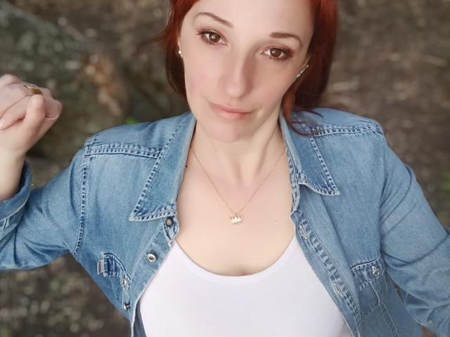 MissieLorrie, com cabelo ruivo, vestindo uma jaqueta jeans azul e uma camiseta branca, mostrando seus seios, com a mão direita levantada, olhando para a câmera com um leve sorriso..