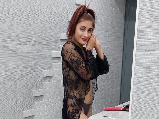 Marina, sexy y atractiva, con una coleta roja, una blusa negra de encaje transparente que deja ver los pezones, una vulva pequeña, de pie frente a una pared de ladrillos blancos, sonriendo, con las manos cerca de la cara, un pincel de maquillaje rojo sobre un borde..