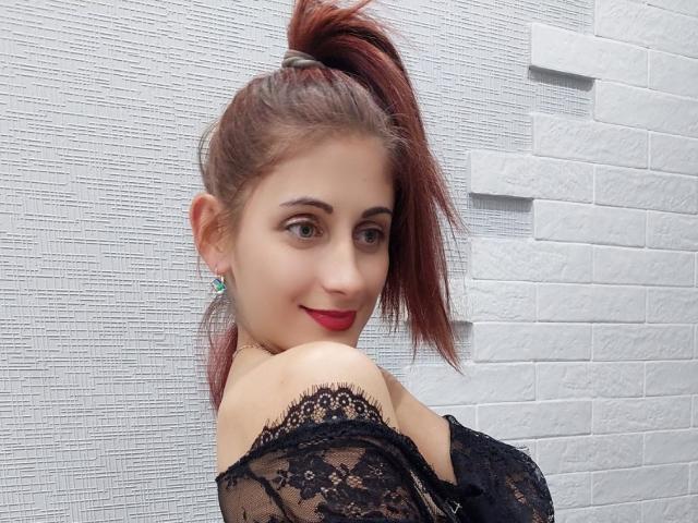 Marina, sexy en aantrekkelijk, met rood haar in een staart, een zwarte kanten top met open schouders, rode lippenstift en turquoise oorbellen, lacht naar de camera en laat haar borsten zien..