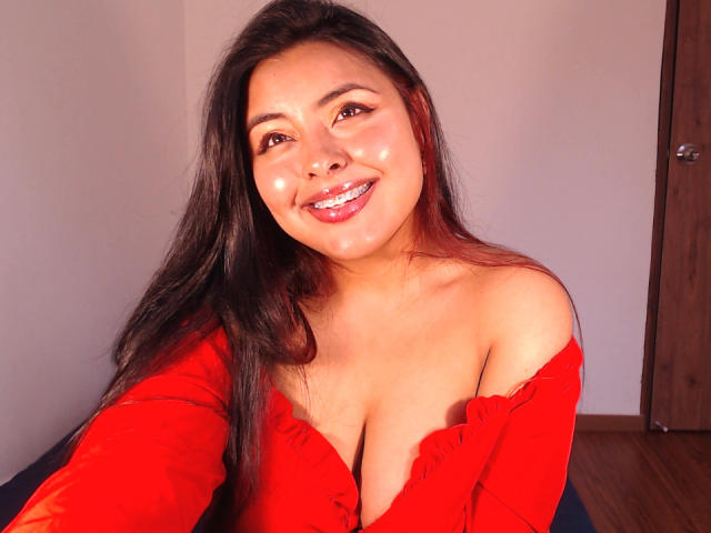 AngelaRoggers with long dark hair, smiling, wearing a red off-shoulder top, exposing her big boobs, shiny face, and red lipstick, standing in a wooden room.