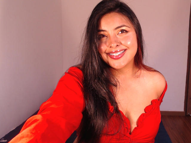 Angela Roggers, con cabello negro largo, sonriendo, vestida con una blusa roja con un hombro descubierto, mostrando sus pequeños senos, y una sustancia blanca en la cara, tomando un selfie..