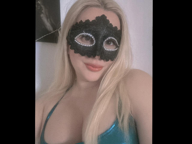 RoseStaar, com cabelo loiro, usando uma máscara preta com glitter e olhos de diamante, e uma blusa verde brilhante que mostra seus seios grandes. Ela está sorrindo com batom laranja..