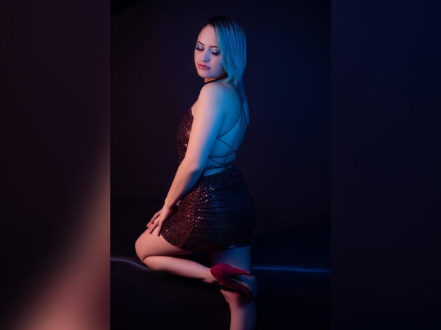 Cassie Jones, con i capelli biondi, indossa un abito nero scintillante, tacchi alti rossi, e mostra il sedere, è inginocchiata in un ambiente con scarsa illuminazione, e guarda oltre la spalla con un leggero sorriso..