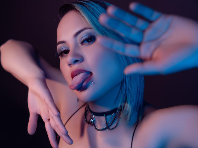 CassieJones with blue-tinted face, tongue out, tongue piercing, blonde hair, black choker, chain necklace, and hands in frame, showing off her sexy, edgy look.