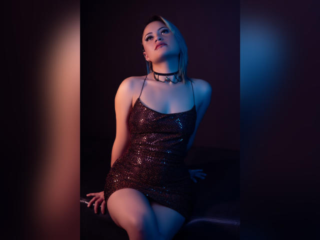 Cassie Jones, vêtue d'une robe noire scintillante, avec un collier et des cheveux blonds courts, assise sur un canapé noir, les jambes jointes, regardant vers le haut avec les lèvres légèrement entrouvertes, dans une lumière bleue et rose tamisée..