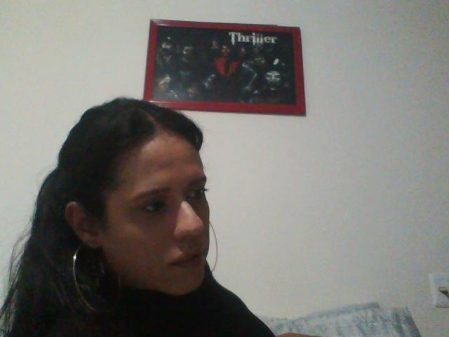Andressa69 mit dunklem Haar, Reifenohrringen und ernstem Gesichtsausdruck, trägt ein schwarzes Oberteil und befindet sich in einem Raum mit einem "Thinner"-Poster an einer weißen Wand..