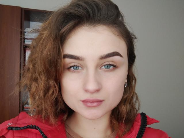 LisaViv a les yeux bleus, les cheveux bruns ondulés, et porte une veste rouge. Ses sourcils sont épais et elle a du rouge à lèvres rose. Elle regarde directement l'appareil photo..
