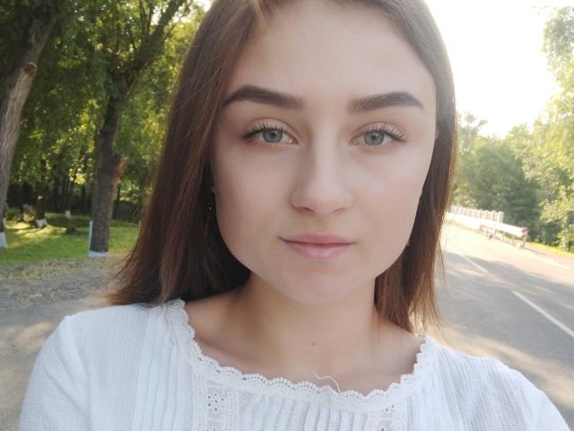 LisaViv, con ojos verde claro, cabello castaño, una blusa blanca de encaje y una sonrisa sutil, mostrando su pequeña nariz y labios carnosos, de pie en una calle soleada..